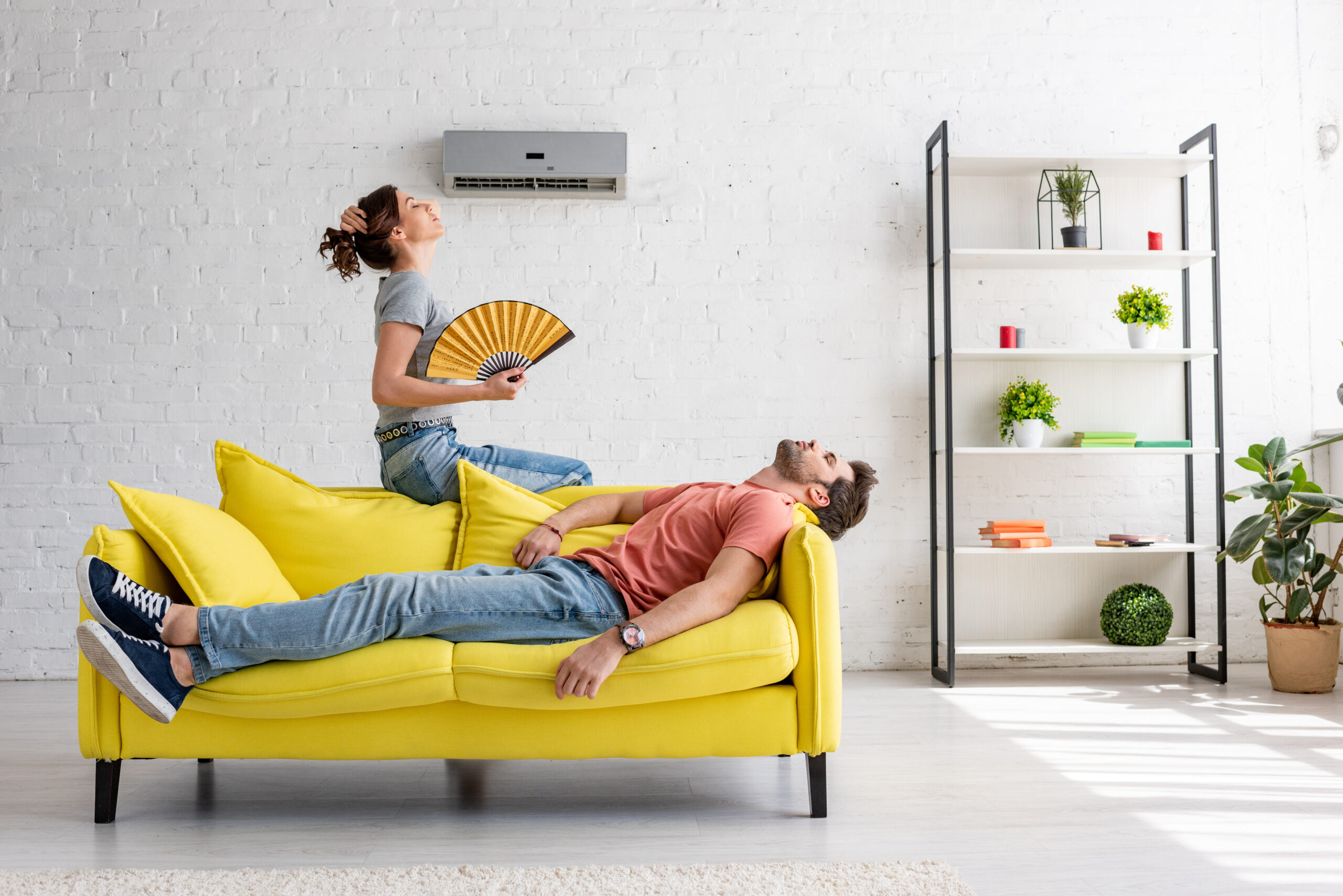 homme épuisé allongé sur un canapé jaune sous un climatiseur près d'une femme avec un ventilateur à main
